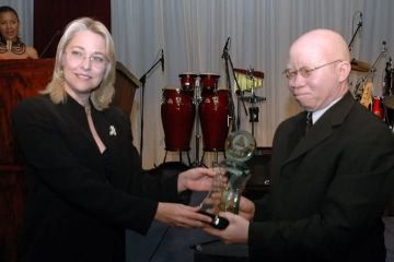 A woman in a black suit presents a glass award to a man with albinism wearing a dark suit, with musical instruments visible onstage behind them.
