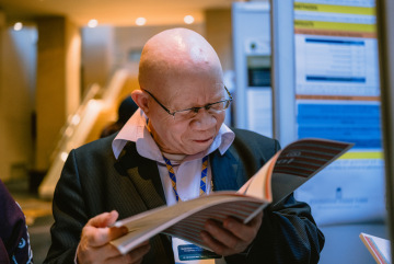 Patrick Wadula wearing glasses and a dark suit reads an open conference publication at an indoor event, with information displays visible in the background.