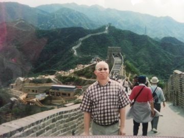 A person with albinism wearing sunglasses stands on the Great Wall of China, with green mountains and other visitors in the background.