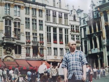 A person with albinism wearing sunglasses stands in a busy historic city square lined with ornate buildings.